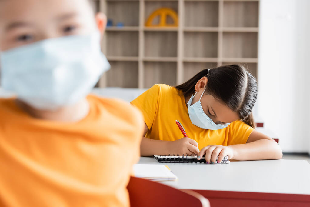 Ασιάτισσα μαθήτρια με ιατρική μάσκα που γράφει στο σημειωματάριο κατά τη διάρκεια μαθήματος - Φωτογραφία, εικόνα
