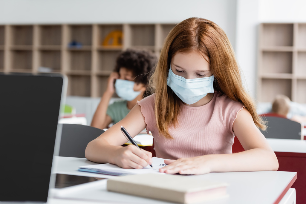 ruda uczennica w masce medycznej pisanie w notatniku podczas lekcji - Zdjęcie, obraz