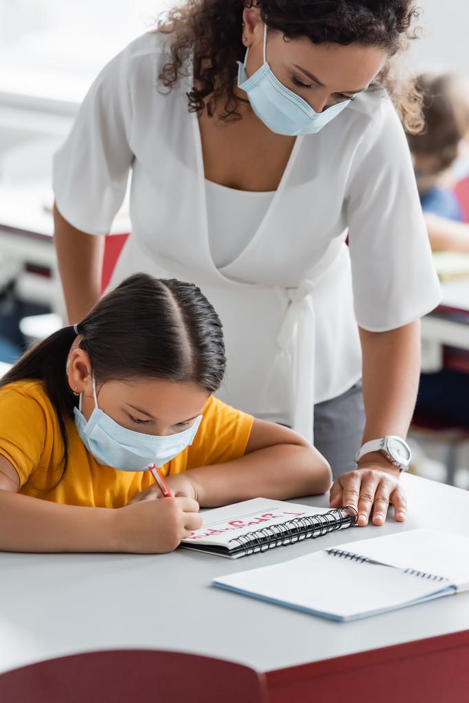 africano americano insegnante in medico maschera in piedi vicino asiatico ragazza scrittura in notebook - Foto, immagini