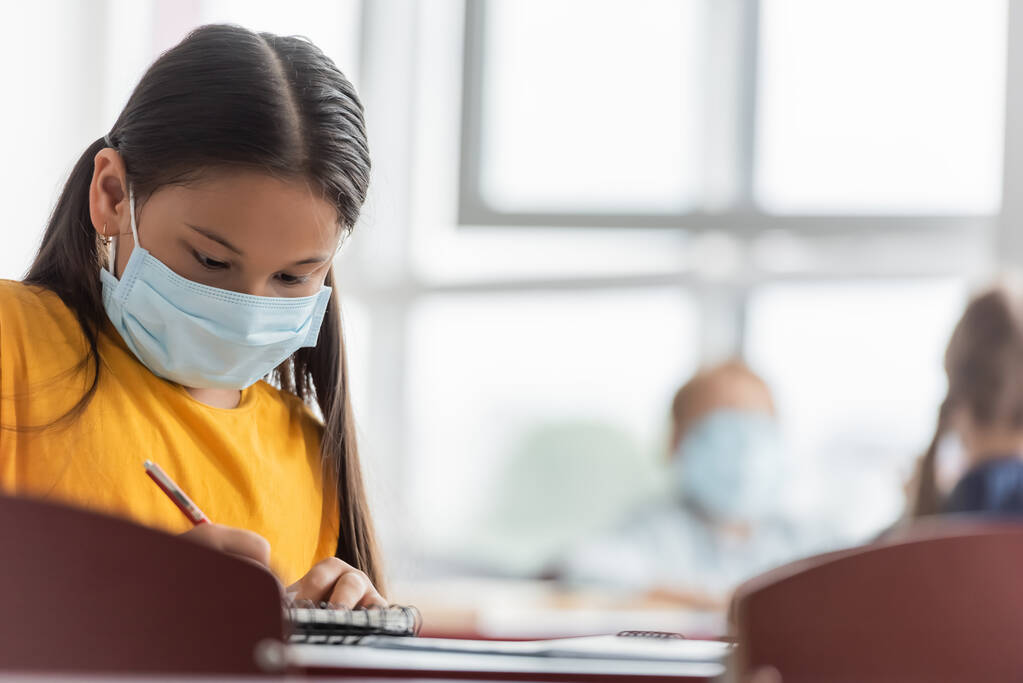 asiático colegiala en médico máscara escritura en notebook en aula - Foto, imagen