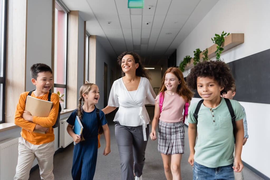 enfants multiculturels joyeux marchant dans le couloir de l'école avec professeur afro-américain - Photo, image