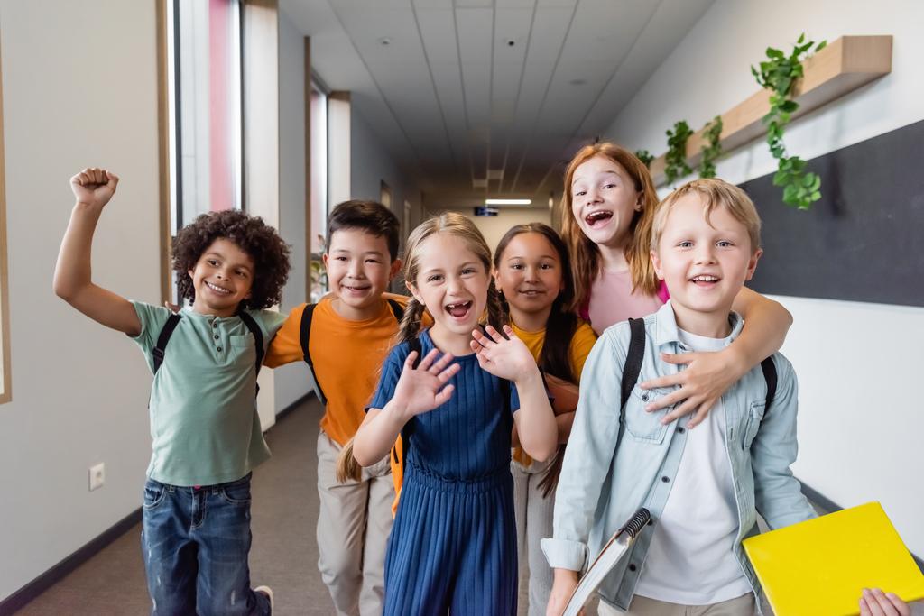radosne wieloetniczne dzieci obejmując, machając rękami i pokazując radosny gest w szkole - Zdjęcie, obraz
