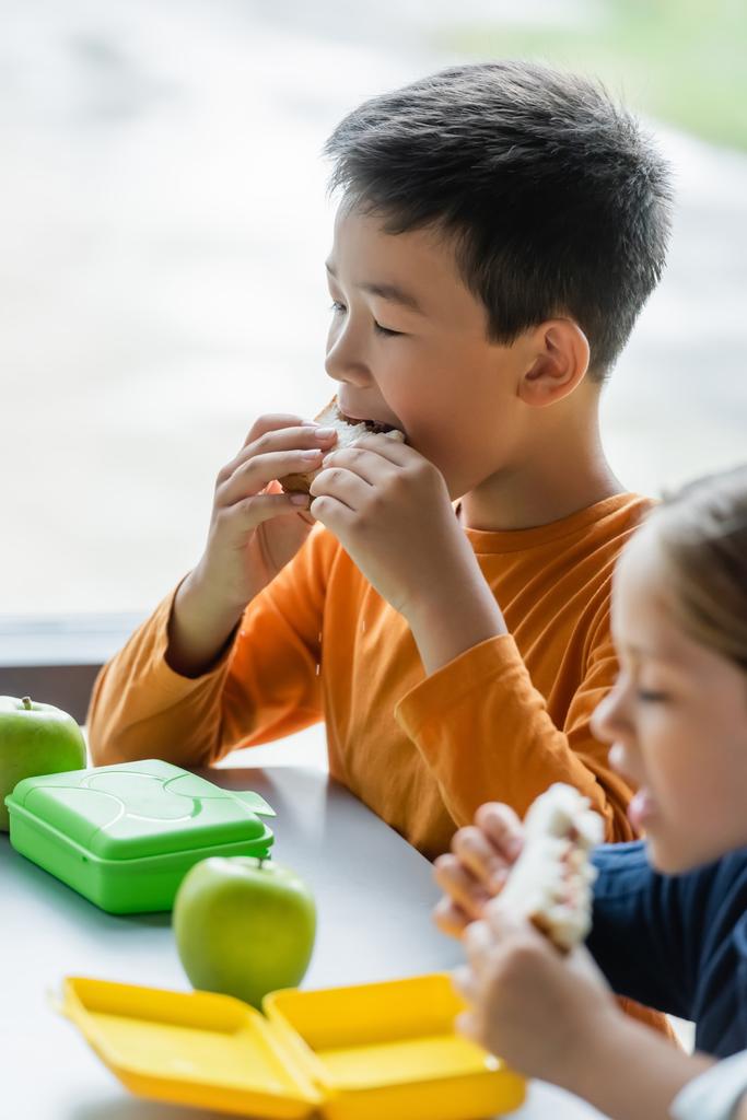 asian schoolboy eating sandwich near blurred girl in school eatery - Photo, Image