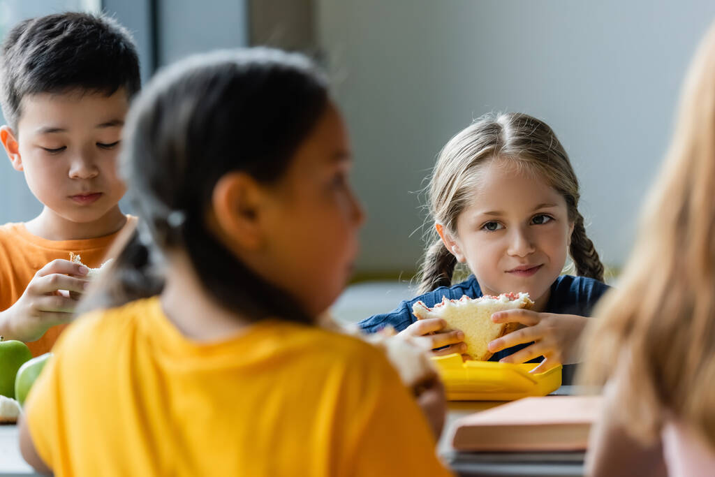 школярка їсть бутерброд біля розмитих азіатських друзів
  - Фото, зображення