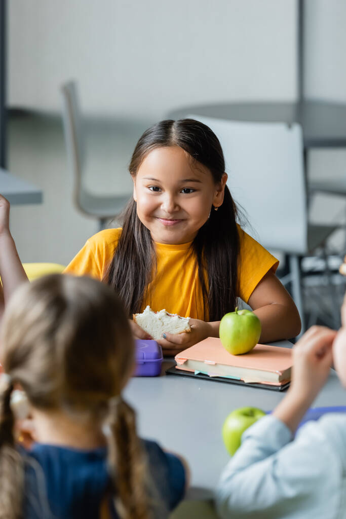 щаслива азіатська школярка тримає сендвіч під час обіду з розмитими однокласниками
 - Фото, зображення