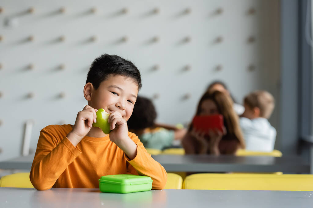 alegre asiático chico con fresco manzana sonriendo en cámara en escuela eatery - Foto, imagen