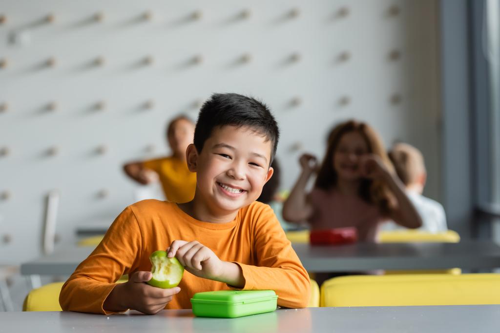 feliz asiático niño celebración con fresco manzana sonriendo en cámara cerca borrosa niños en escuela cantina - Foto, imagen