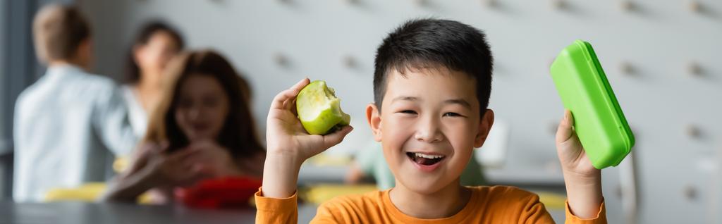 wesoły azjatycki chłopiec trzyma pudełko na lunch i jabłko w pobliżu rozmazane dzieci w szkolnej jadłodajni, baner - Zdjęcie, obraz