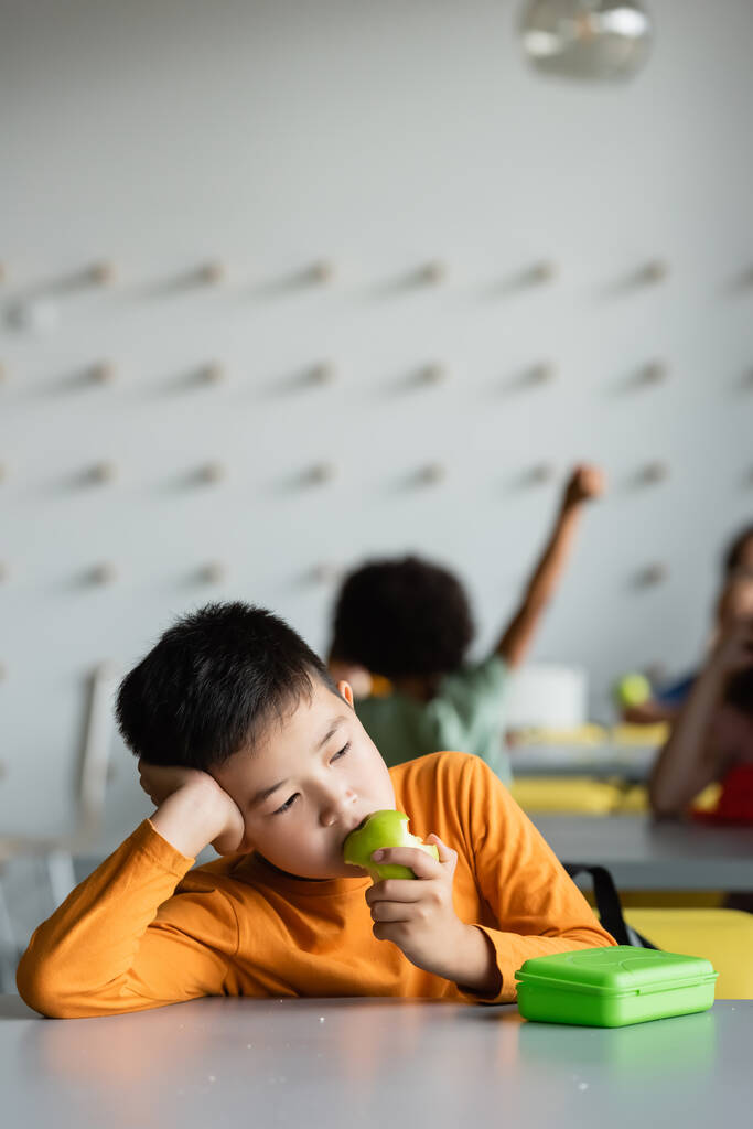 surullinen aasialainen poika syö omenaa koulussa ruokapaikka lähellä lapsia hämärtynyt tausta - Valokuva, kuva
