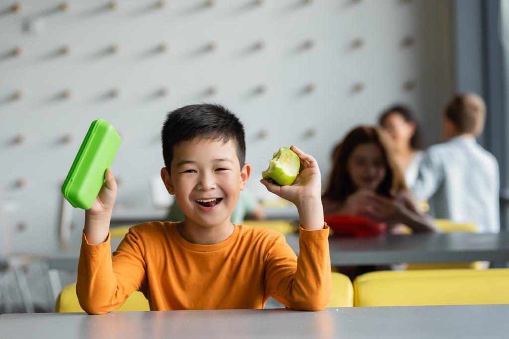 radosny Azji chłopiec trzyma lunch pudełko i jabłko w szkoła jadalnia  - Zdjęcie, obraz