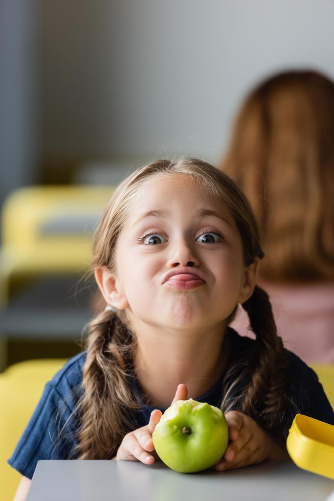 játékos lány kidugja a nyelvét, miközben grimaszol közel alma az iskolai menzán - Fotó, kép