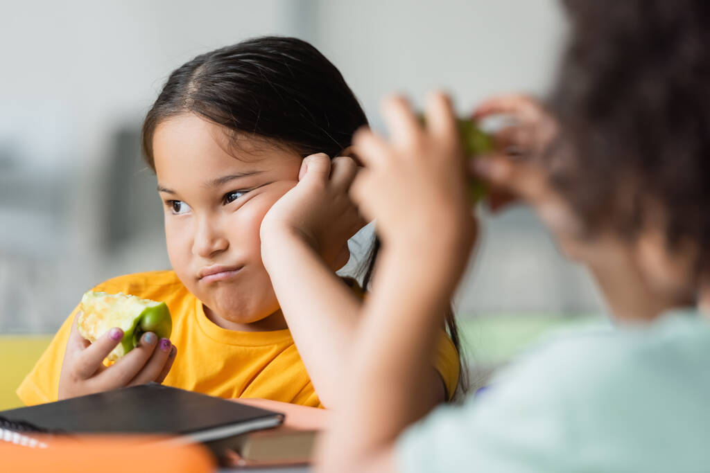 disgustada chica asiática con manzana cerca borrosa africano americano chico en escuela cantina - Foto, imagen