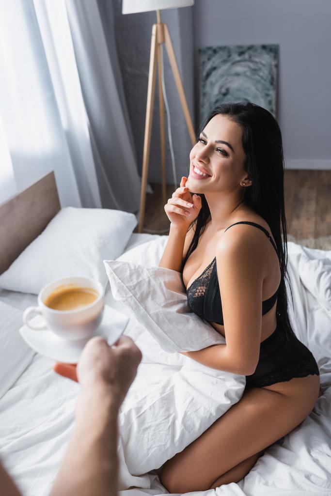 verschwommener Mann hält Tasse Cappuccino neben fröhlicher sexy Frau in Unterwäsche - Foto, Bild