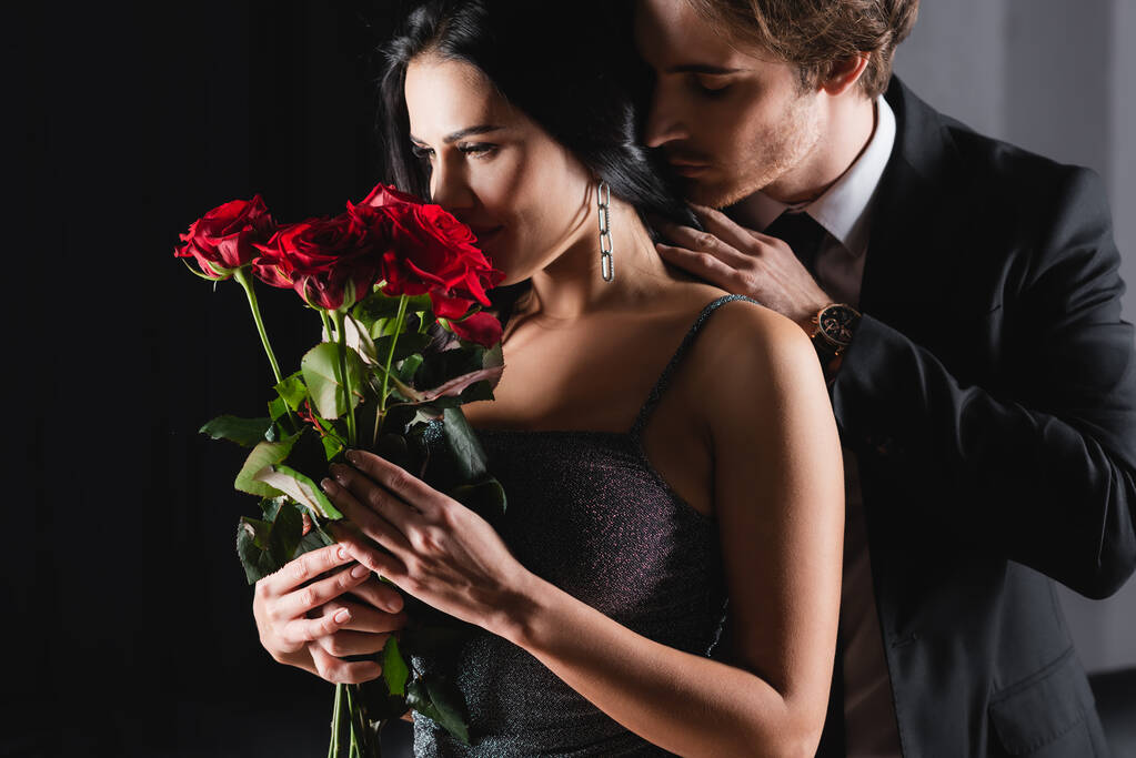 joven mujer oliendo ramo de rosas rojas cerca de hombre en traje en negro  - Foto, imagen