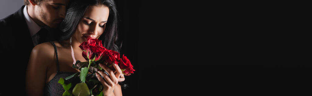 sonriente mujer sosteniendo rosas cerca de novio en negro, bandera - Foto, Imagen