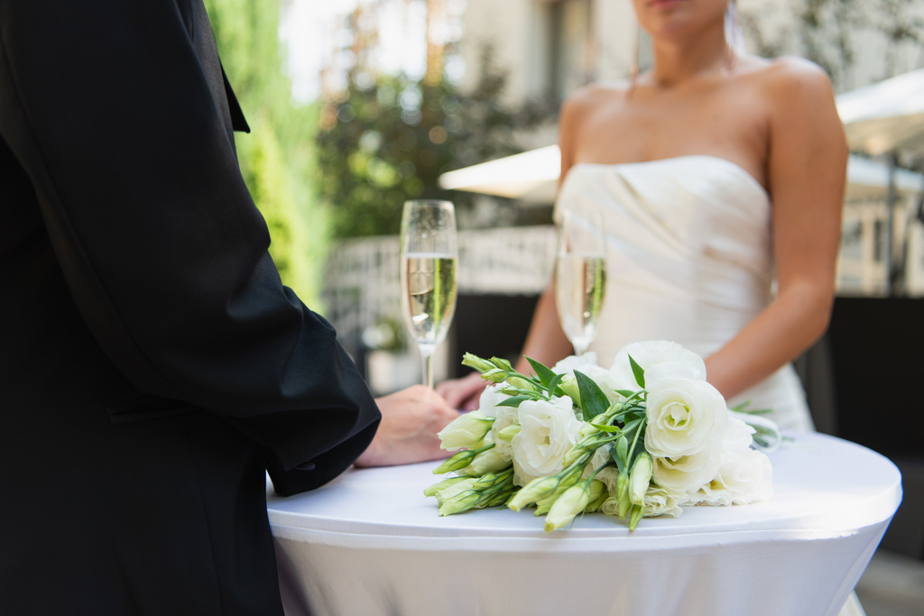 Vista ritagliata della donna lesbica in abito in piedi vicino al bouquet di nozze, champagne e fidanzata all'aperto  - Foto, immagini