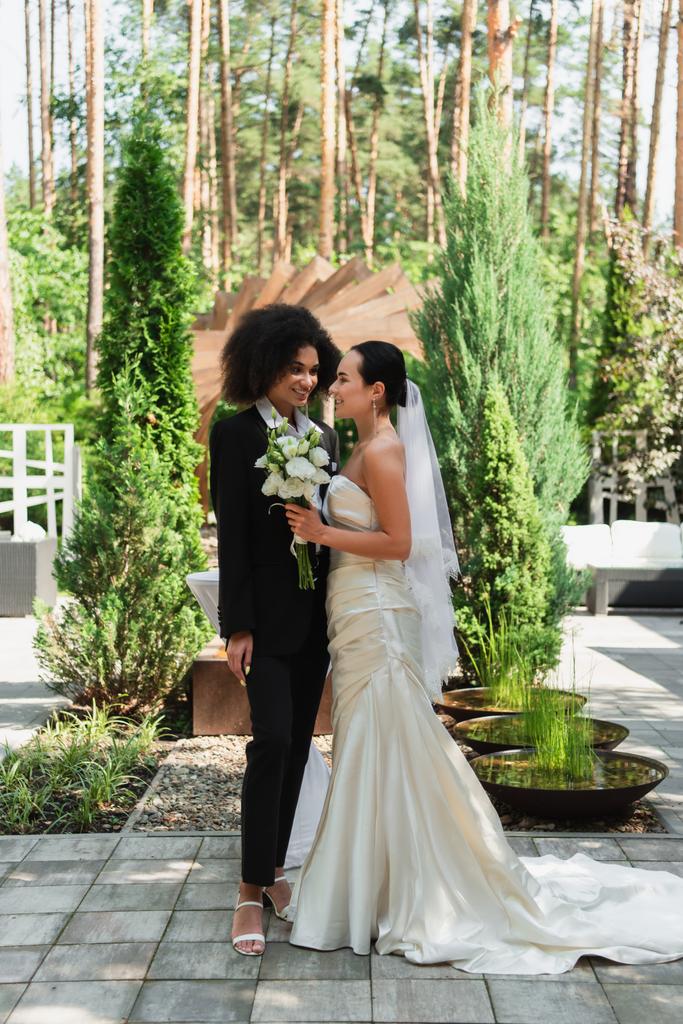 Alegre interracial lesbianas pareja con ramo de mirar el uno al otro durante la boda al aire libre  - Foto, Imagen