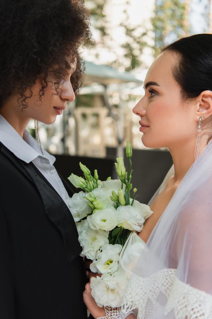 Vista lateral de mulheres lésbicas inter-raciais com buquê olhando uns para os outros durante o casamento ao ar livre  - Foto, Imagem