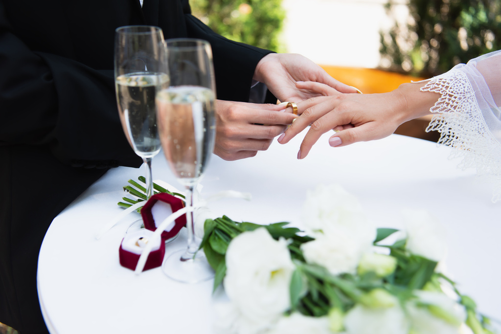 Ausgeschnittene Ansicht einer lesbischen Frau, die Ehering am Finger ihrer Freundin in der Nähe von Champagner und Strauß trägt  - Foto, Bild