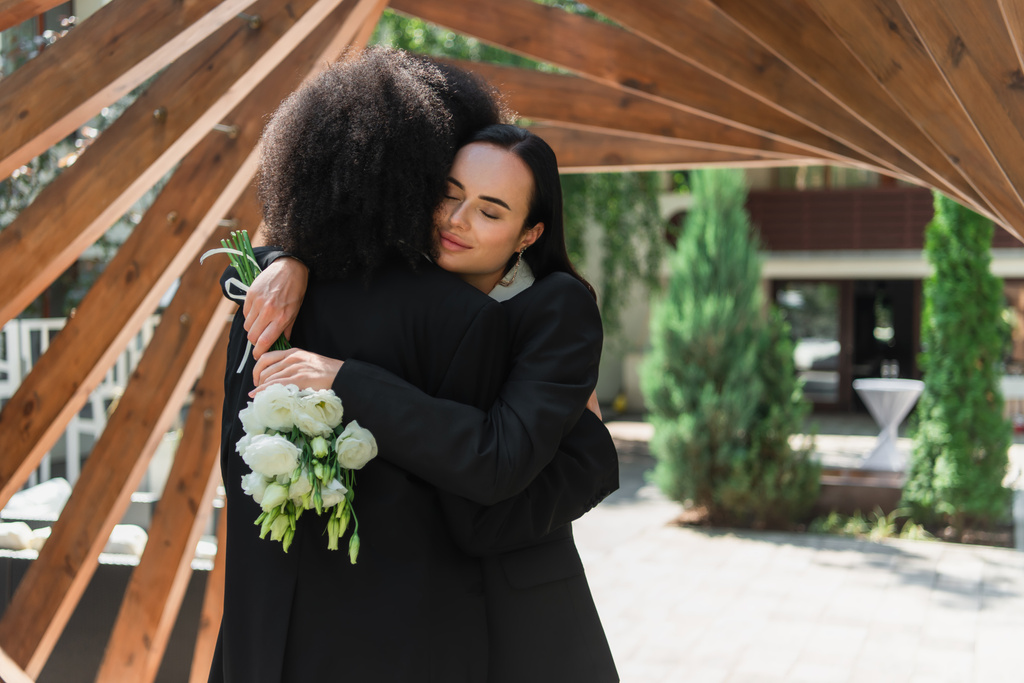 Lesbian woman with wedding bouquet hugging african american girlfriend under arch in park  - Photo, Image