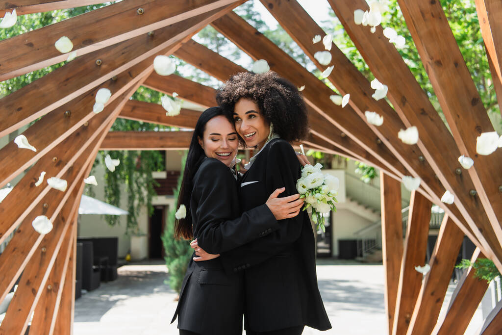 Parkta gül yapraklarının altında düğün buketi ile sarılan mutlu ırklar arası lezbiyen çift.  - Fotoğraf, Görsel