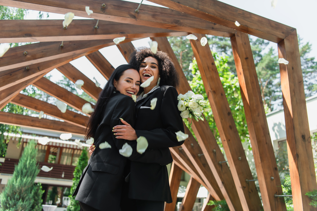 alegre multiétnica lesbiana pareja en trajes abrazando cerca pétalos durante la boda en parque  - Foto, Imagen
