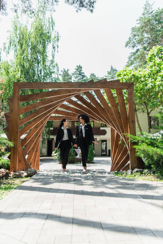Улыбающаяся многонациональная лесбийская пара в костюмах со свадебным букетом гуляющая в парке  - Фото, изображение