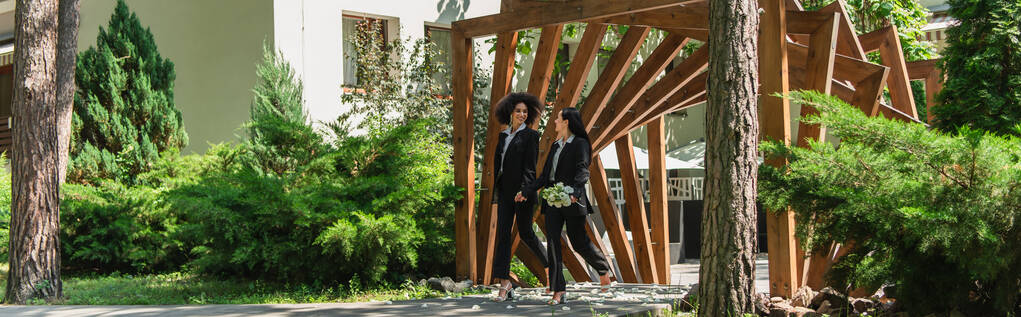 Multiethnisches gleichgeschlechtliches Paar mit Hochzeitsstrauß zu Fuß im Park, Banner  - Foto, Bild