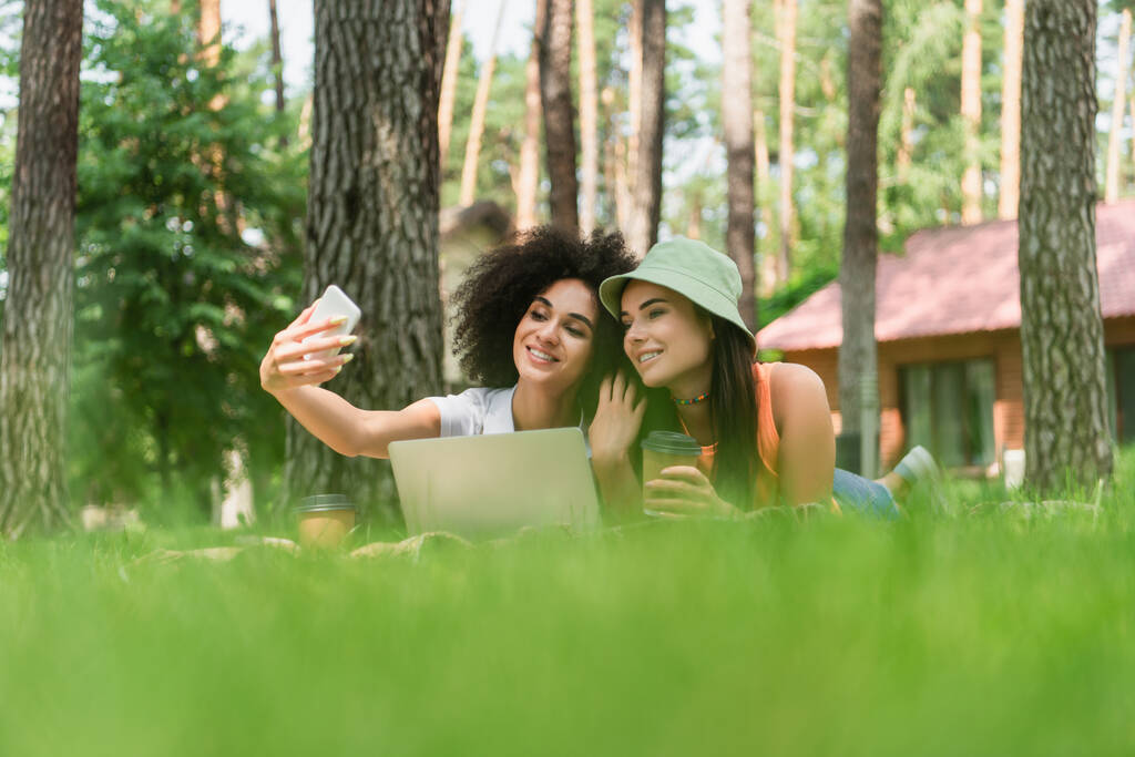 Donna afroamericana che prende selfie vicino alla ragazza con caffè e laptop nel parco  - Foto, immagini