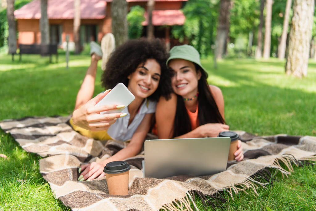 Blurred interracial same sex couple taking selfie near laptop and coffee in park  - Photo, Image