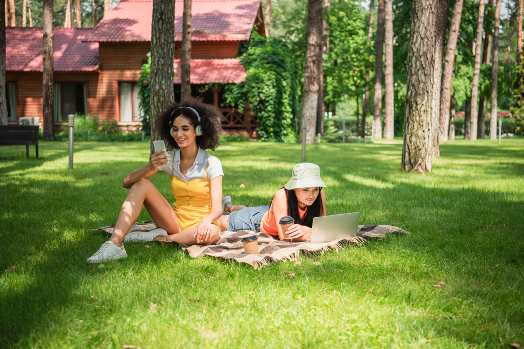 Afroamerykanka w słuchawkach za pomocą smartfona w pobliżu dziewczyny z kawą i laptopem na trawniku  - Zdjęcie, obraz