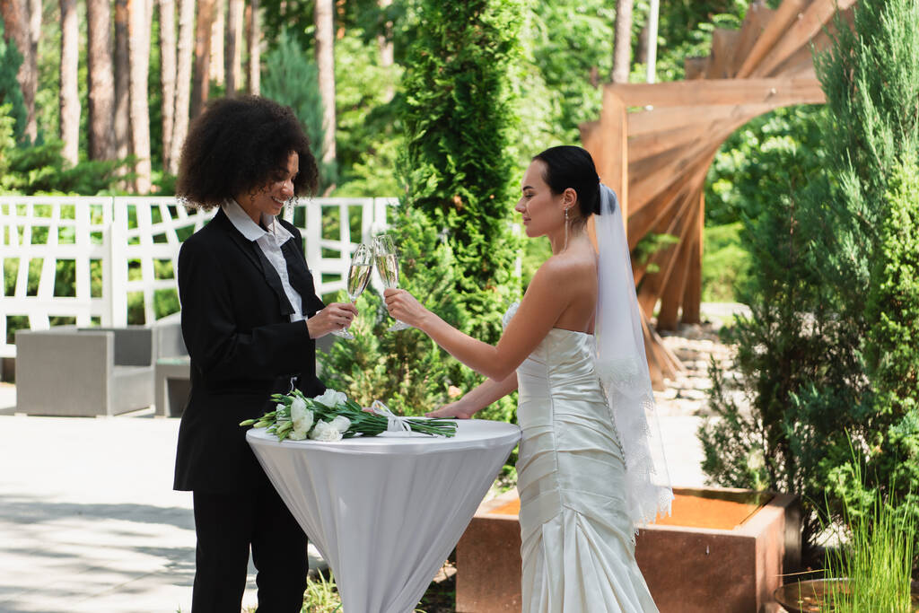 alegre interracial casal clinking com champanhe durante casamento ao ar livre  - Foto, Imagem