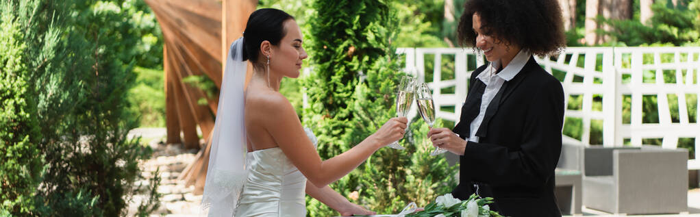 pozytywna międzyrasowa lesbijska para brzęcząca z szampanem podczas wesela na świeżym powietrzu, baner  - Zdjęcie, obraz