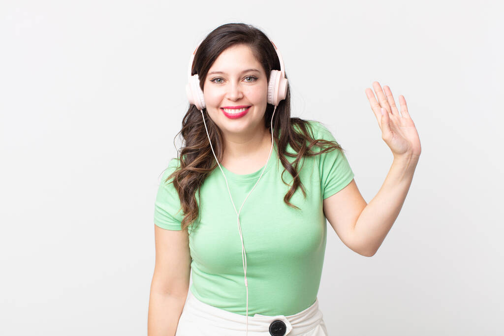 hübsche Frau lächelt glücklich, winkt mit der Hand, begrüßt und begrüßt Sie beim Musikhören mit Kopfhörern - Foto, Bild
