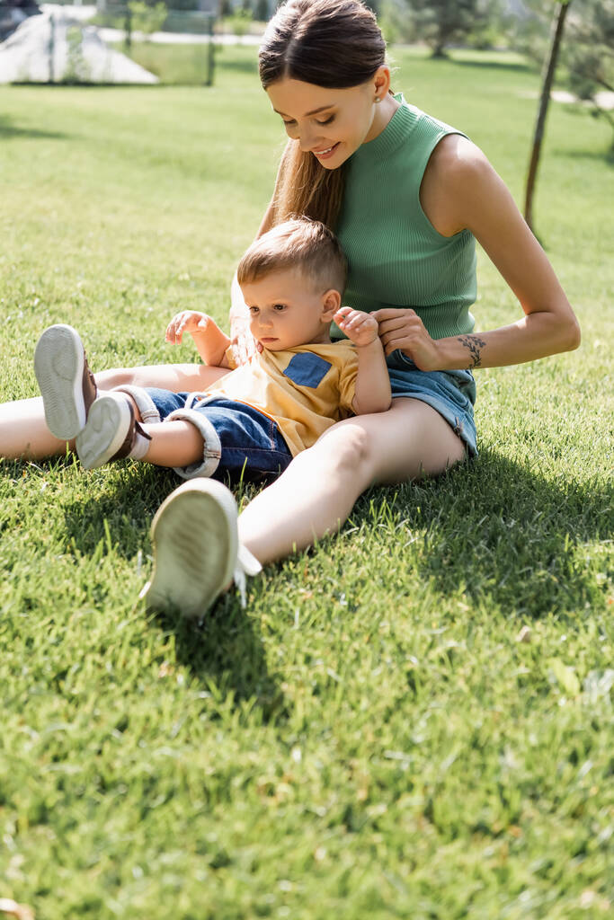 alegre madre sentada en la hierba con su hijo pequeño fuera  - Foto, imagen