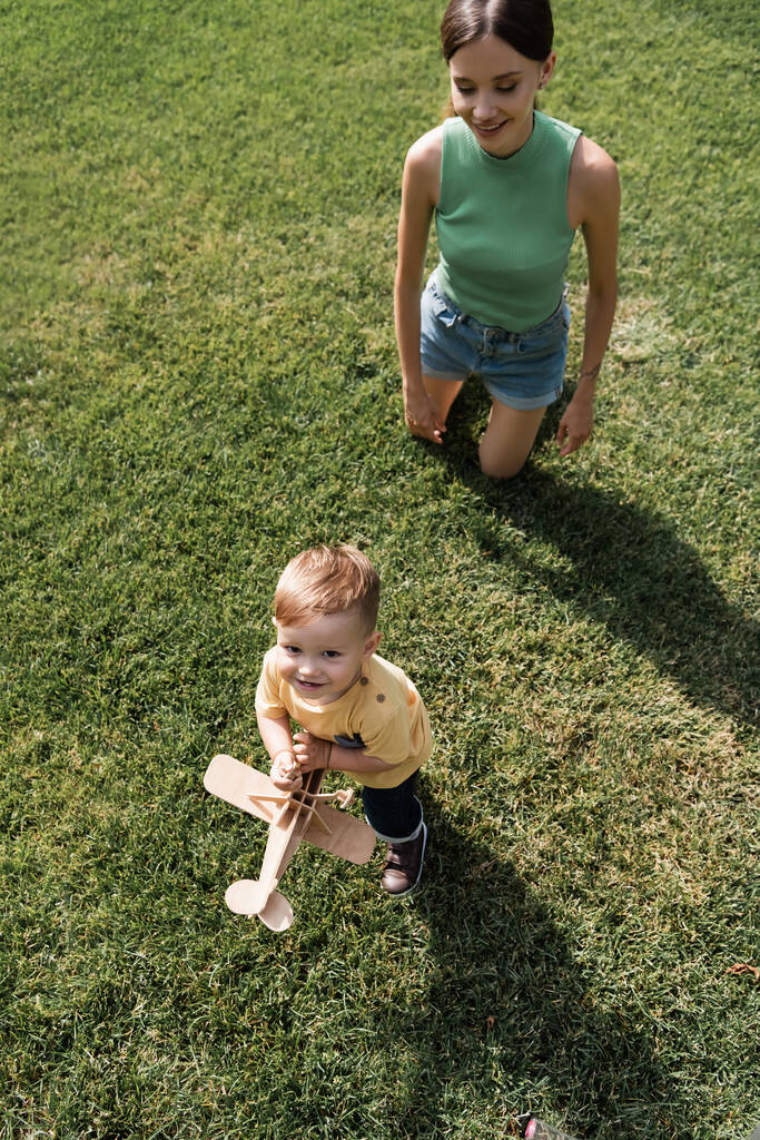 Ansicht von oben: Glückliche junge Mutter schaut fröhlichen Kleinkind-Sohn mit Spielzeug-Doppeldecker an und steht auf Gras - Foto, Bild