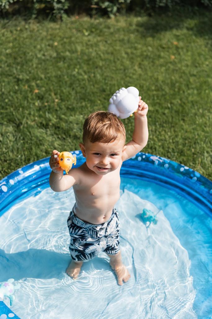 high angle view of happy toddler boy in swim trunks playing with rubber toys in inflatable pool outside  - Photo, Image