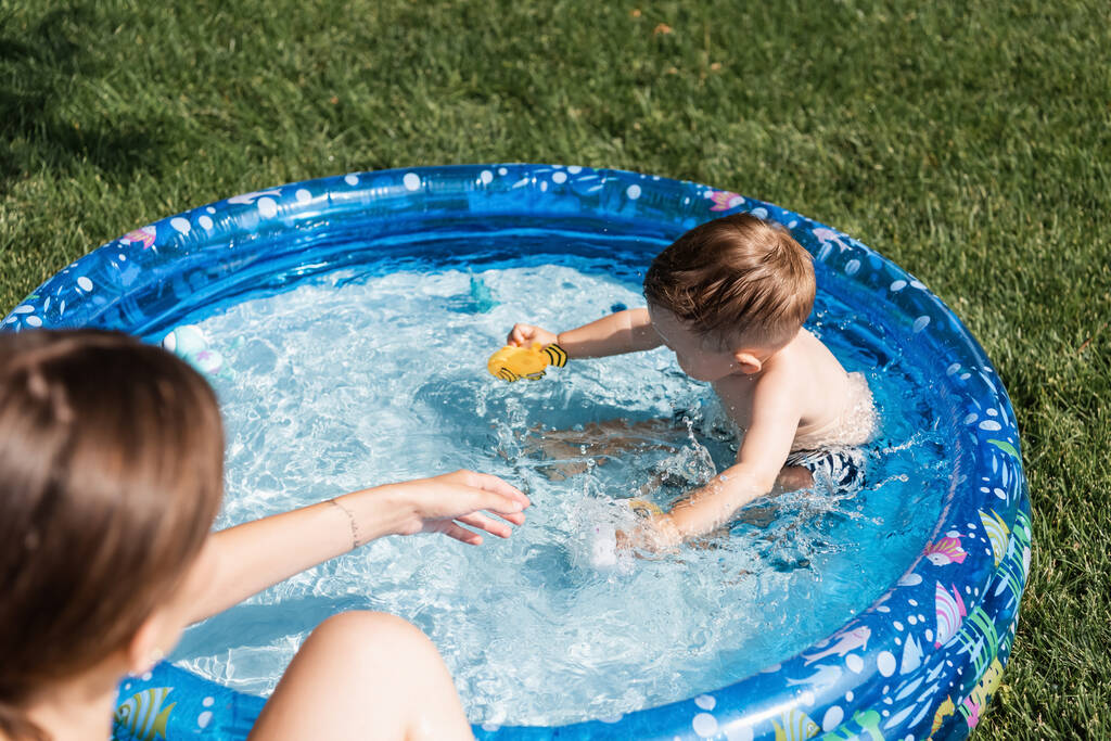 high angle view of toddler boy playing with rubber toys in inflatable pool near blurred mother  - Photo, Image