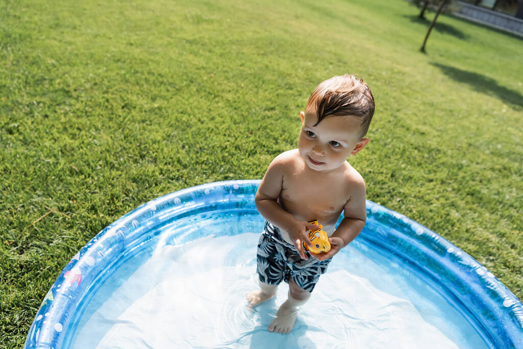 hoge hoek uitzicht op peuter kind in zwembroek staan in opblaasbaar zwembad met rubber speelgoed - Foto, afbeelding