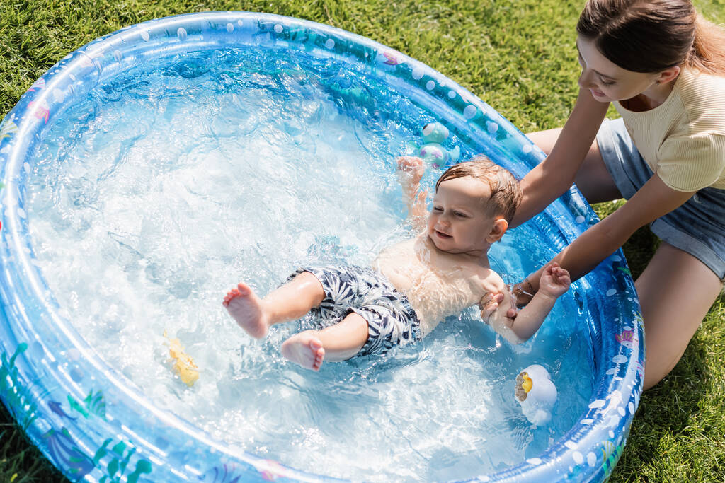 visão de ângulo alto da mãe sorridente que banha o filho sem camisa da criança na piscina inflável - Foto, Imagem