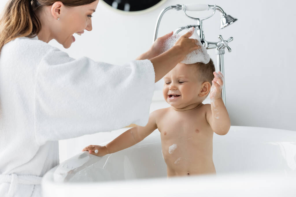 Bornozlu neşeli anne küvette banyo yapan küçük çocuk.  - Fotoğraf, Görsel
