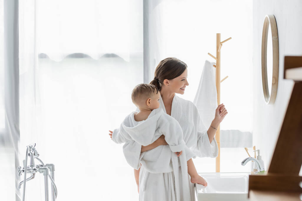 Seitenansicht einer fröhlichen Mutter, die ihren kleinen Sohn im Arm hält und im Badezimmer auf den Spiegel schaut  - Foto, Bild