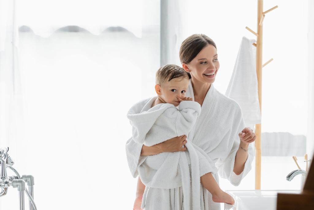 весела мати тримає в руках маленького сина і дивиться у ванну кімнату
 - Фото, зображення
