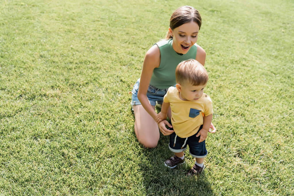 Erstaunte Mutter sieht Kleinkind auf grünem Gras stehen  - Foto, Bild