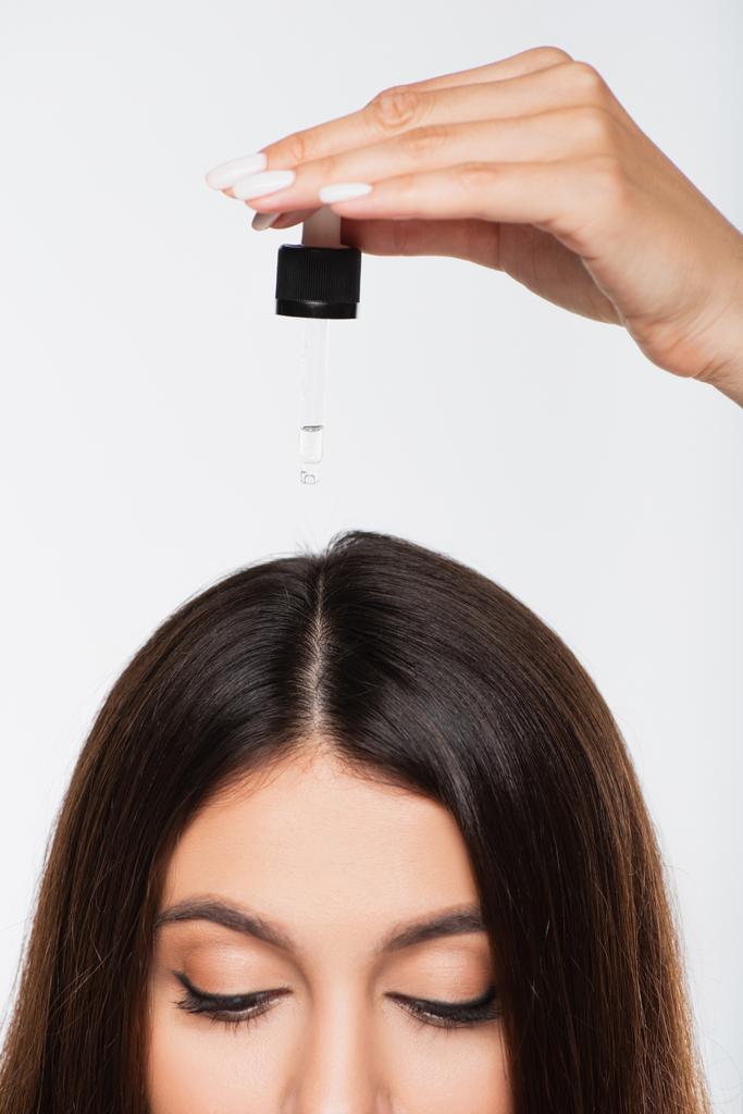 abgeschnittene Ansicht einer jungen Frau mit glänzendem Haar, die Behandlungsöl auf den Kopf aufträgt, isoliert auf grau - Foto, Bild
