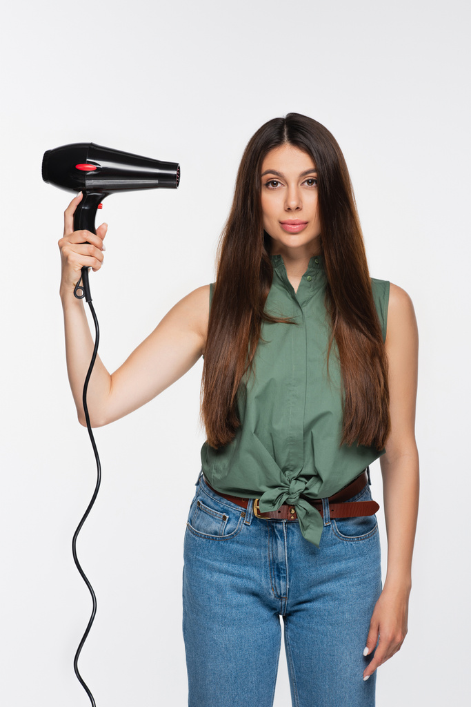 όμορφη νεαρή γυναίκα με λαμπερά μαλλιά κρατώντας στεγνωτήρα μαλλιών απομονώνονται σε γκρι  - Φωτογραφία, εικόνα