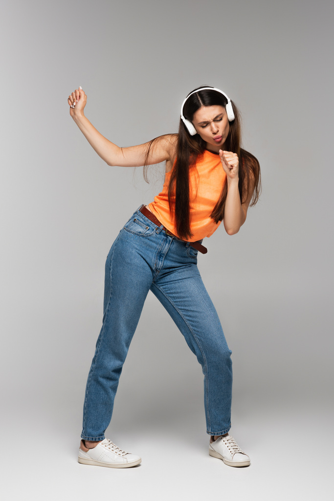 pełna długość brunetka kobieta w dżinsach dżinsy i słuchawki bezprzewodowe słuchanie muzyki podczas tańca na szary - Zdjęcie, obraz
