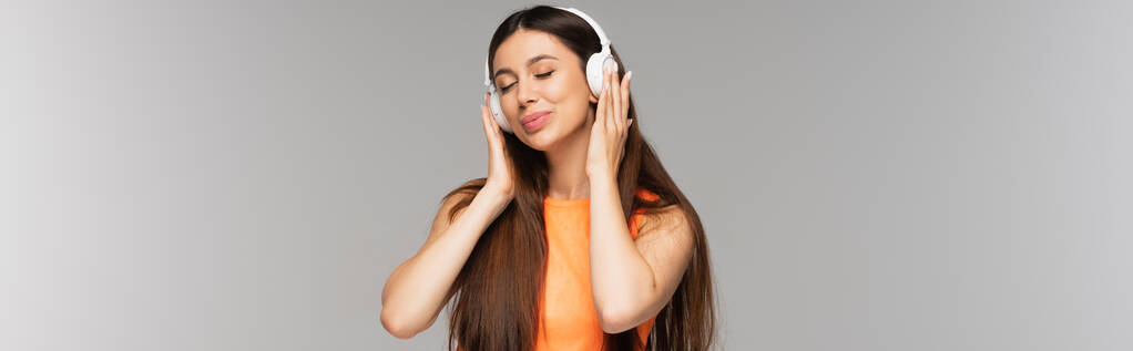 ευχαριστημένη γυναίκα με τζιν τζιν και ασύρματα ακουστικά ακούγοντας μουσική και χαμογελώντας απομονωμένη σε γκρι, banner - Φωτογραφία, εικόνα