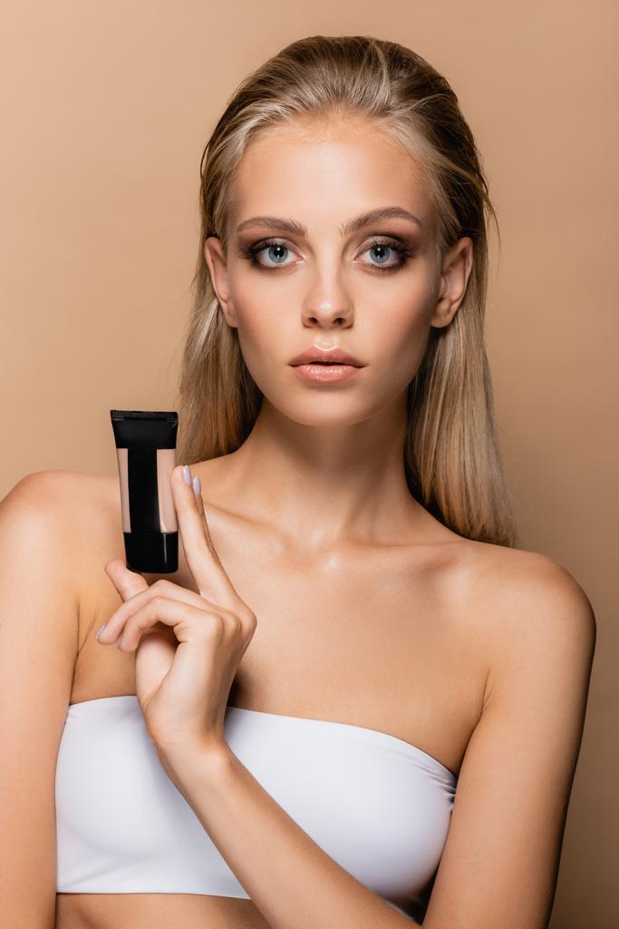 pretty blonde woman with grey eyes showing tube with face foundation isolated on beige - Photo, Image