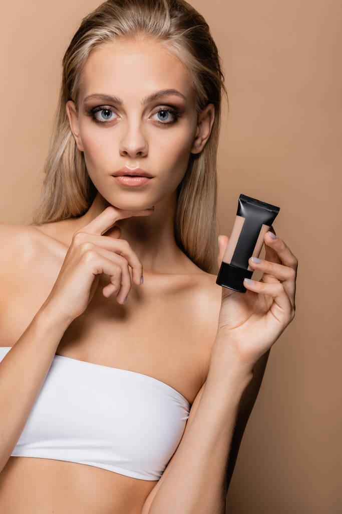 young woman with perfect skin holding face foundation while touching chin isolated on beige - Photo, Image